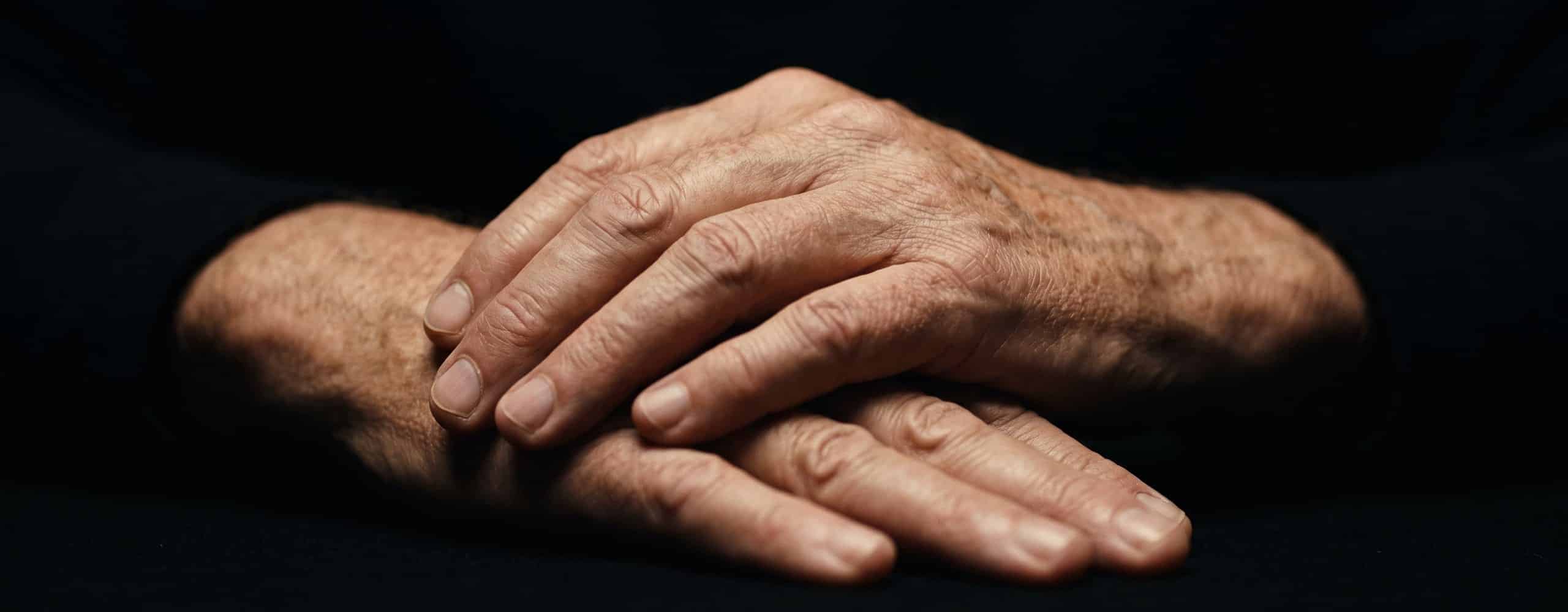 Mains de personne âgée posées l'une sur l'autre sur fond sombre, relief des articulations éclairé par une lumière rasante